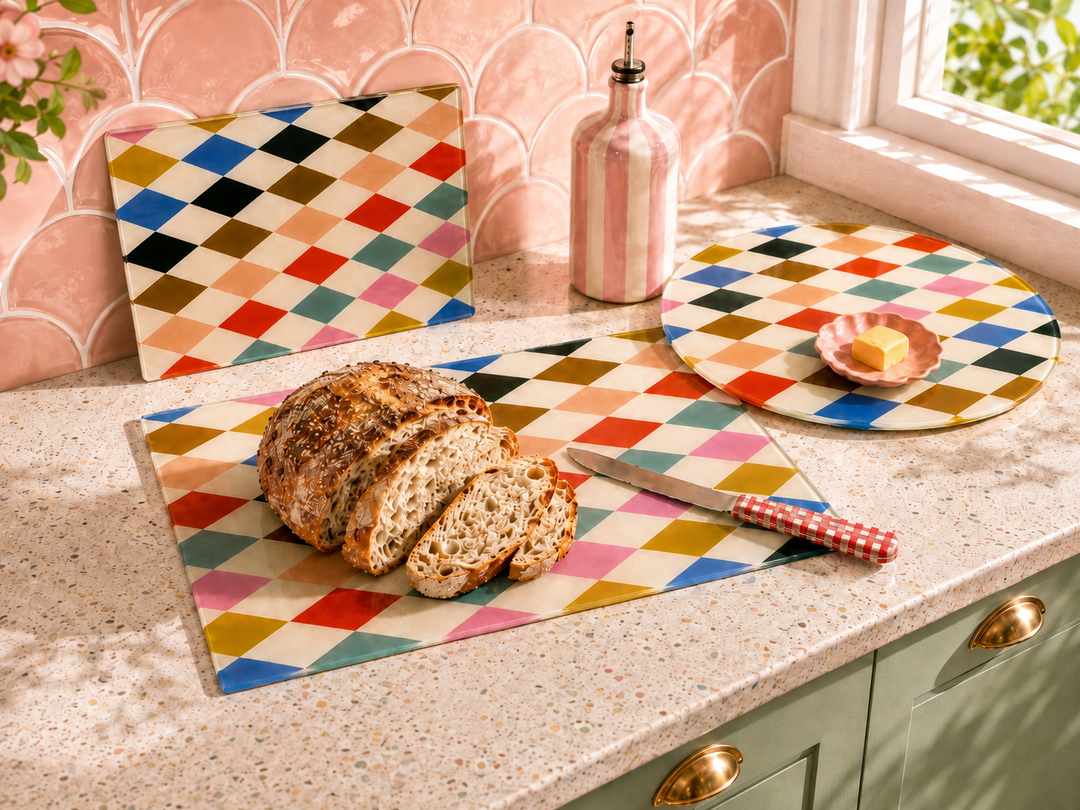 Three Yililo glass chopping boards in the Patch Pop Diamond design on a kitchen worktop: the small 20 x 28cm board leaning against pink tiles, the large 28.5 x 39cm board with a sliced loaf, and the 30cm round board.