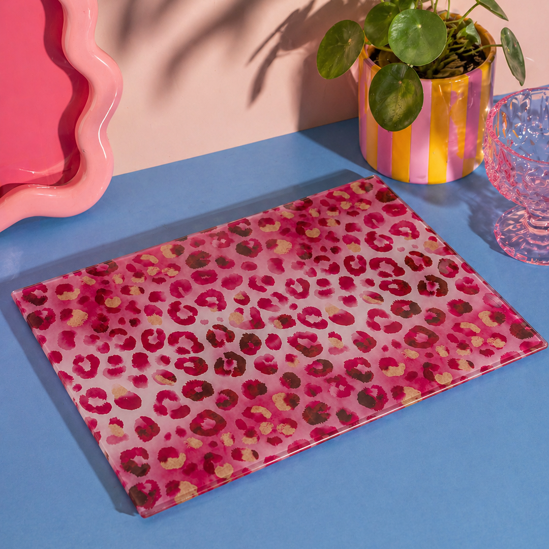 Pink leopard print glass chopping board on a blue surface with a pink wall and plant in the background