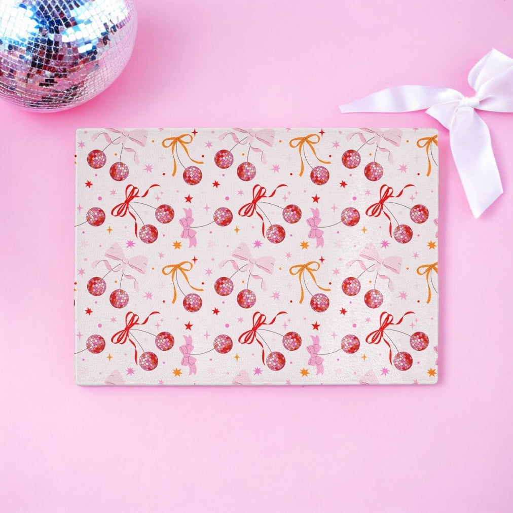 A rectangular sheet with a pink, festive pattern of red disco balls, gold stars, and bows lies on a pink surface. Next to it is Yililos Cherry Disco Bow Coquette Glass Chopping Board and white ribbon, completing the cheerful scene.