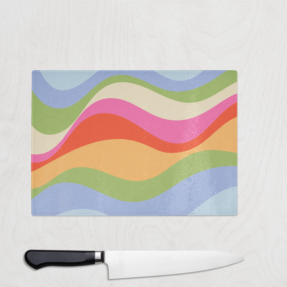 The Yililo Groovy Wavy Glass Chopping Board, featuring blue, green, pink, red, orange, and beige stripes, rests on a light wood surface with a black-handled chef’s knife below this retro kitchen decor piece.