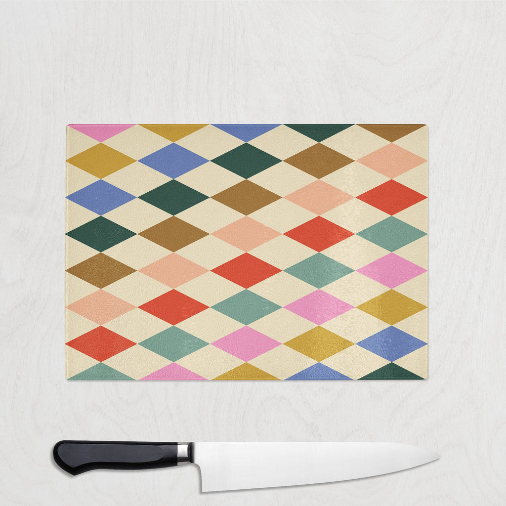 A Yililo Patch Pop Diamond Glass Chopping Board, featuring a vibrant diamond pattern, adds harlequin charm to the light wood surface, while a kitchen knife with a black handle rests below it.