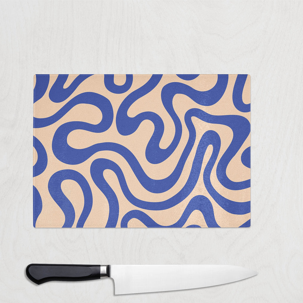 A Yililo Blue Wiggle Funk Glass Chopping Board with a wavy cobalt blue pattern on beige lies on a light surface beside a kitchen knife with a black handle.