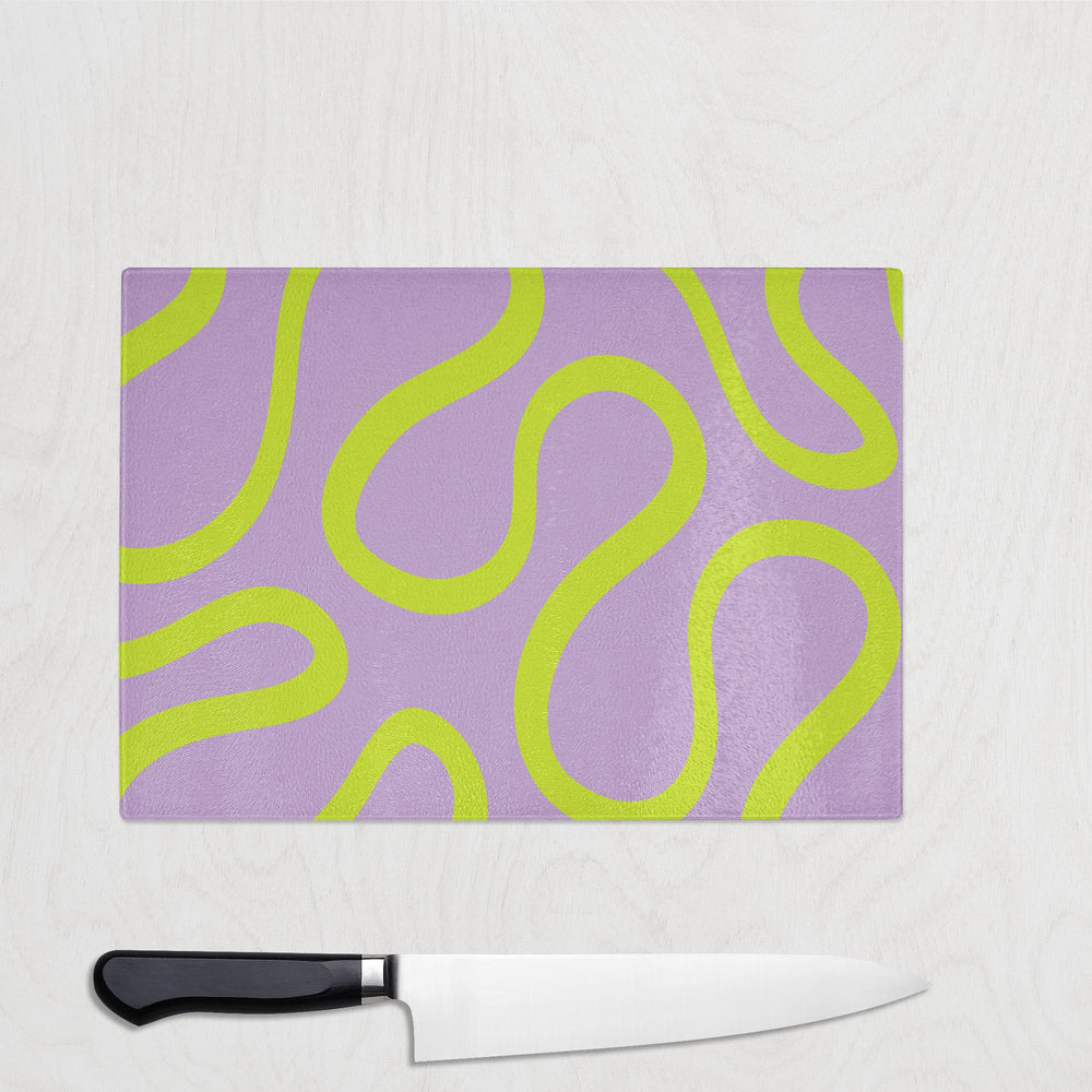 The Yililo Lime Lilac Wiggle Pop Y2K Glass Chopping Board features a wavy lime green pattern on a lavender background, resting on light wood with a large kitchen knife with a black handle placed just below it.