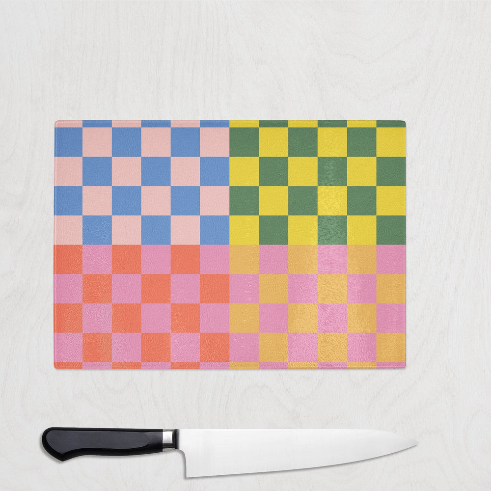 The Yililo Candy Clash Y2K Check Glass Chopping Board, with its vibrant blue, pink, yellow, and green checkered design, brings a playful look to any maximalist kitchen. A black-handled kitchen knife is placed beneath it.