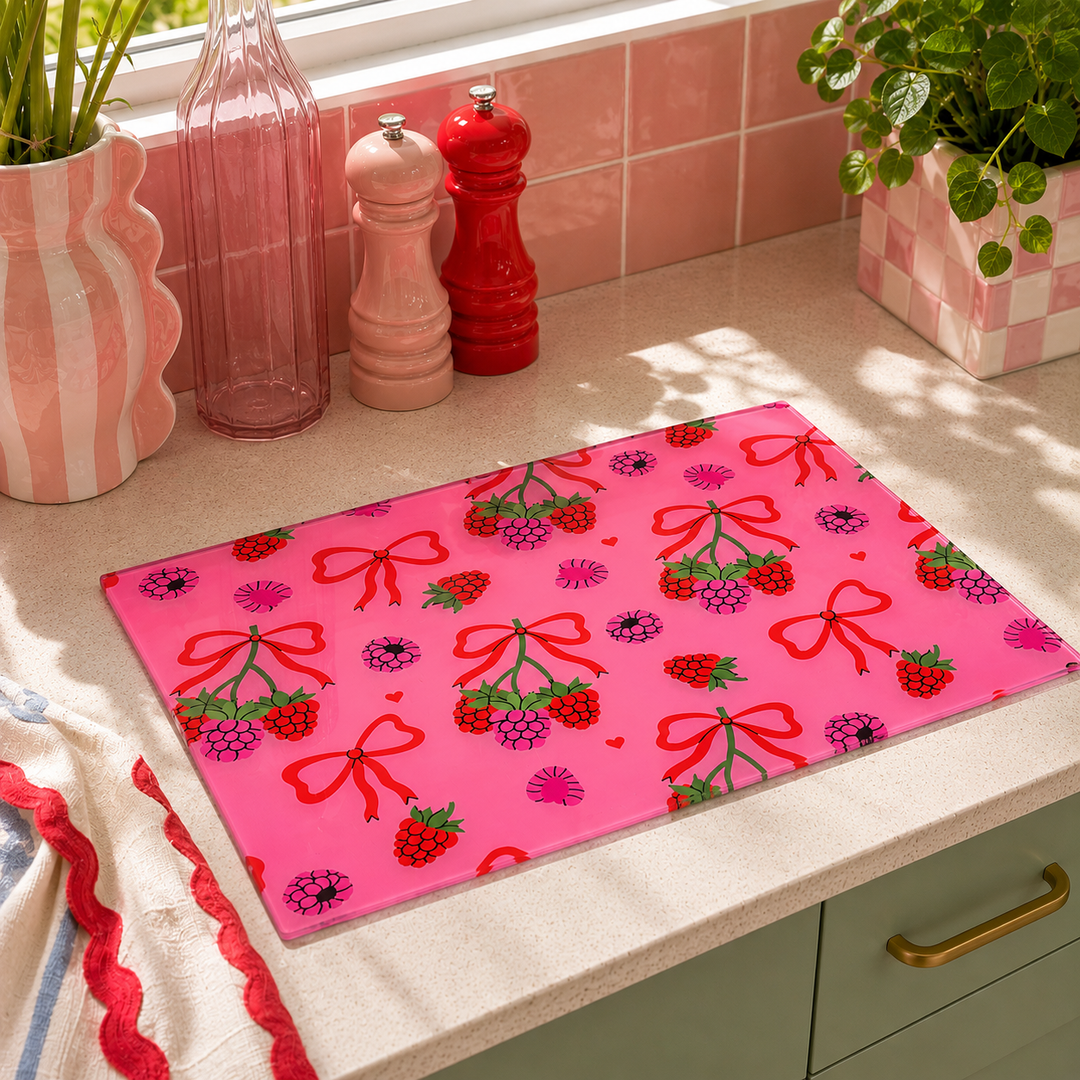 A rectangular pink cutting board with a repeating pattern of red bows, strawberries, and raspberries, resting on a kitchen countertop.