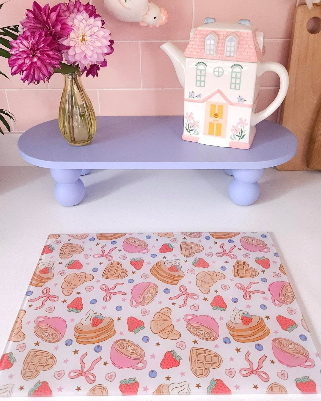 A pastel pink placemat with a whimsical pattern of cakes, pastries, strawberries, and bows is spread on a white table in a kitchen setting. A lavender shelf behind it holds a vase of pink flowers and a decorative ceramic house-shaped teapot, against a pink tiled wall.