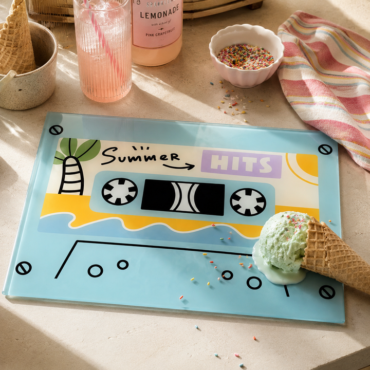 A light blue rectangular glass chopping board with a retro cassette tape design and the words 'Summer Hits,' styled on a beige tabletop with a green ice cream scoop, waffle cones, a pink drink, sprinkles, and a striped cloth.