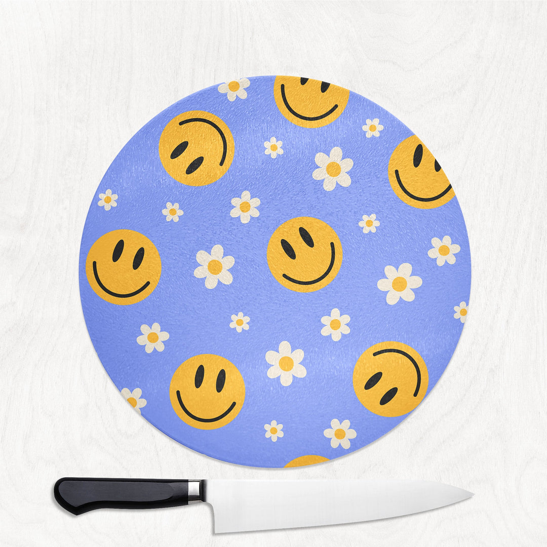 Round blue cutting board with smiley face and flower pattern next to a knife on a white background