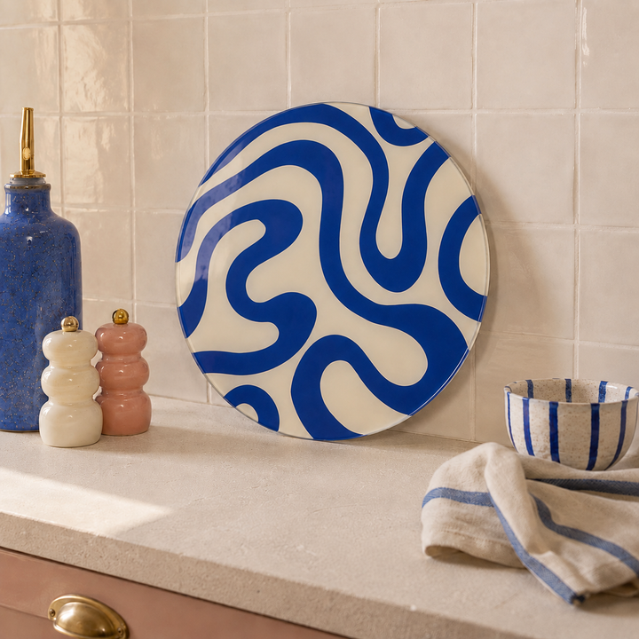 A round glass serving platter with a bold blue wavy pattern on an off-white background, styled on a kitchen counter next to a blue bottle, salt and pepper shakers, a striped bowl, and a striped cloth.