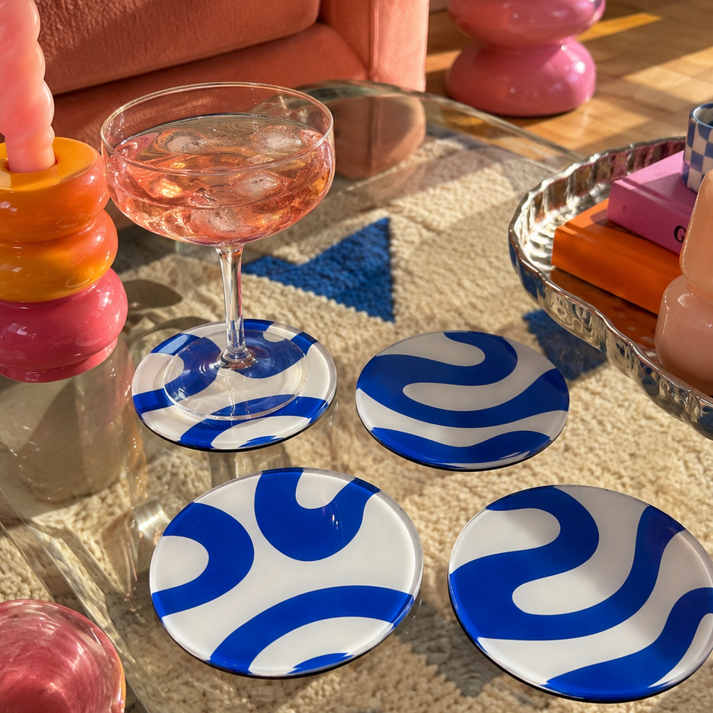 Four round glass coasters with a blue and white wavy abstract pattern arranged on a glass coffee table in a styled living room setting.
