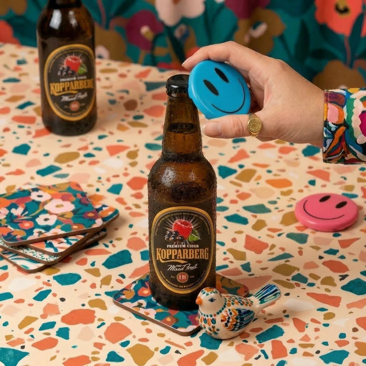 Person opening a Kopparberg cider bottle with a blue bottle opener on a colorful table.