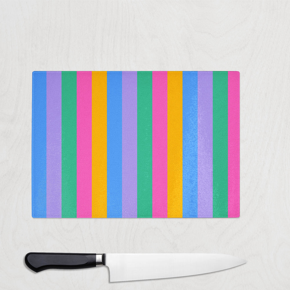 The Yililo Bright Colour Stripe Glass Chopping Board, featuring vertical blue, green, yellow, pink, and purple lines, sits on a light surface with a black-handled kitchen knife below. This stylish board is both heat-resistant and durable.