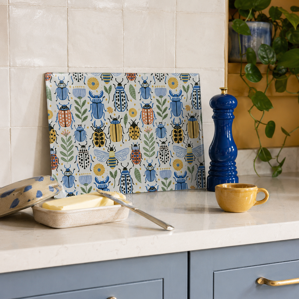A rectangular cutting board with a colourful pattern of illustrated beetles, bees, and flowers on a cream background, leaning on a kitchen counter next to a blue pepper mill, a yellow cup, and a butter dish.