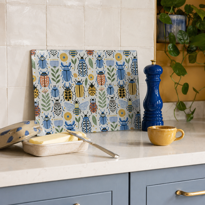 A rectangular cutting board with a colourful pattern of illustrated beetles, bees, and flowers on a cream background, leaning on a kitchen counter next to a blue pepper mill, a yellow cup, and a butter dish.