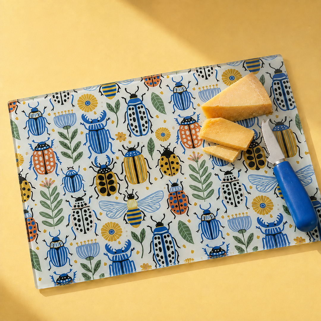 A rectangular cutting board with a colorful illustrated pattern of beetles, bees, flowers, and leaves on a white background, styled with cheese wedges and a blue-handled knife on a yellow surface.