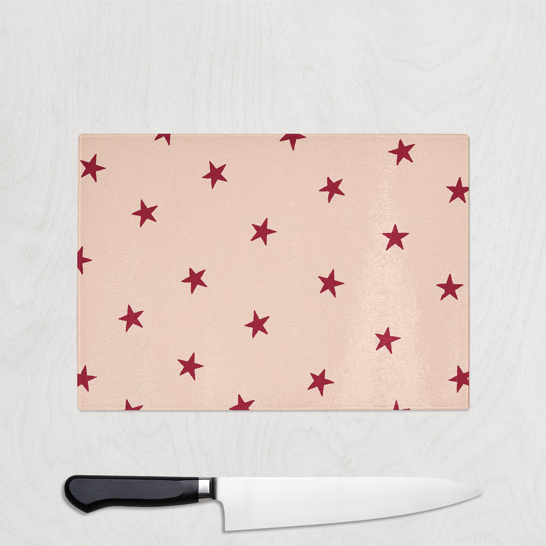 Burgundy Star Christmas glass chopping board with scattered burgundy red stars on a soft blush pink background, shown with a chef's knife