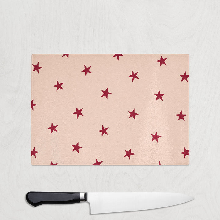 Burgundy Star Christmas glass chopping board with scattered burgundy red stars on a soft blush pink background, shown with a chef's knife