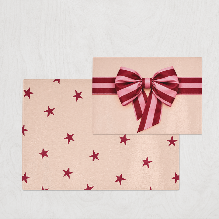 A gift-wrapped style glass chopping board on a light gray textured background. The wrapping paper chopping board is a pale blush pink with scattered dark red stars, and the top rectangular package is tied with a matching pink and dark red striped ribbon and bow.