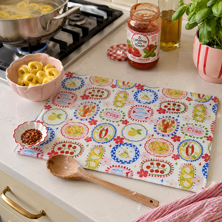 A colourful glass worktop saver with a retro Italian pattern featuring 'Ciao' and food motifs like spaghetti, tomatoes, and peppers, styled on a white countertop with pasta, a wooden spoon, and a jar of tomato sauce.