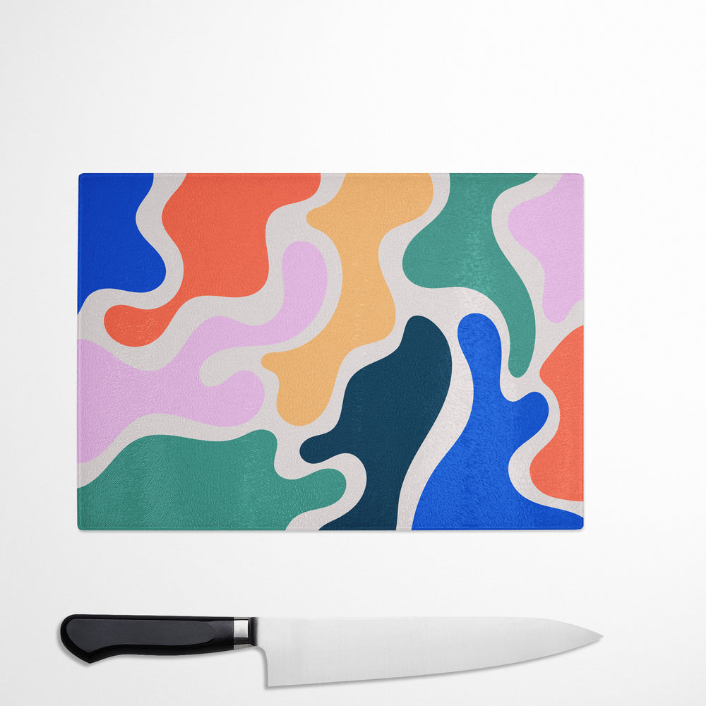 The Yililo Colourful Camouflage Print Glass Chopping Board, featuring vibrant wavy patterns, rests on a white surface beside a black-handled kitchen knife and doubles as a heat-resistant protector.