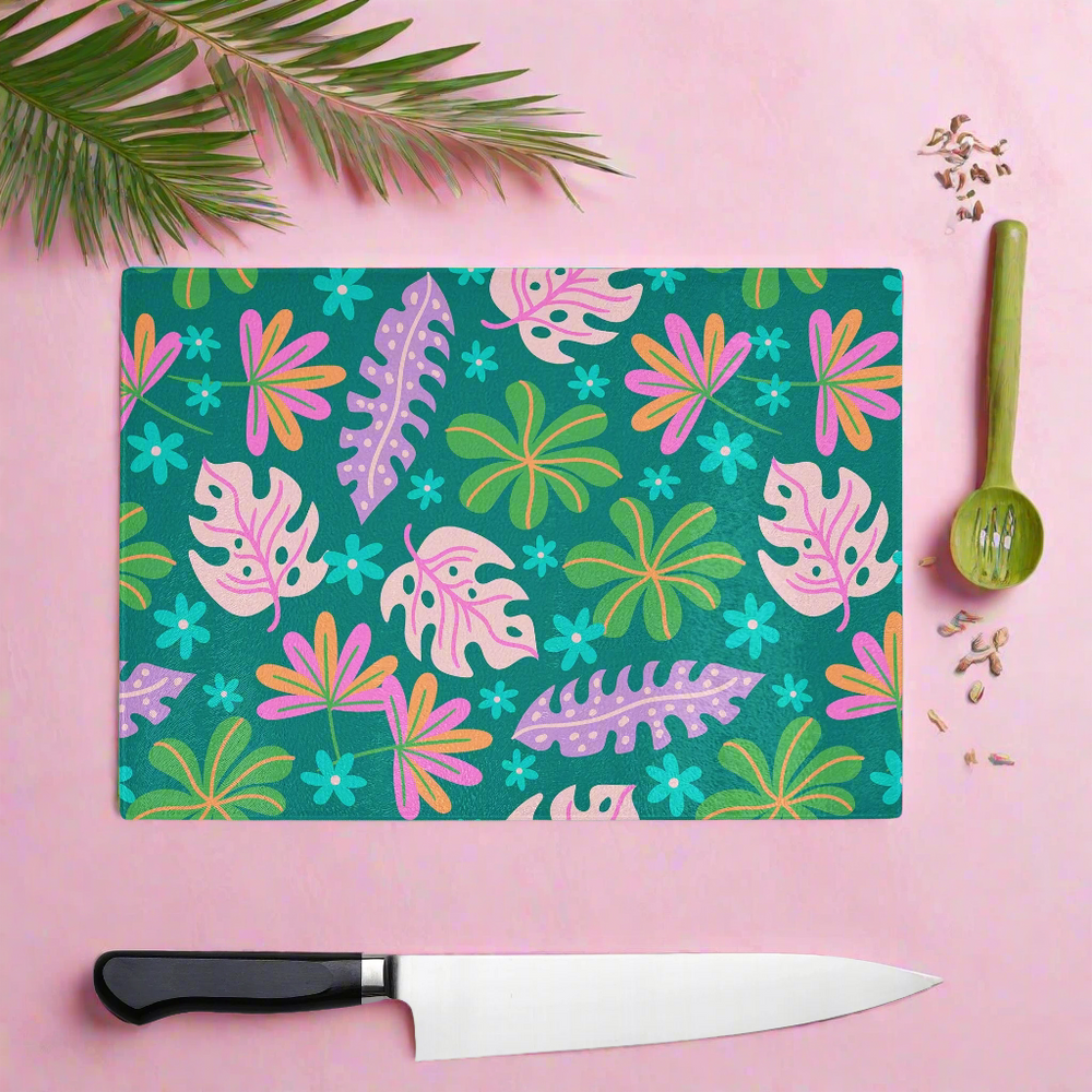 The Yililo Colourful Tropic Print Glass Chopping Board, featuring vibrant leaf and flower designs, rests on a pink surface with a green spoon containing seeds, a black-handled knife, and palm leaves in the top left corner.