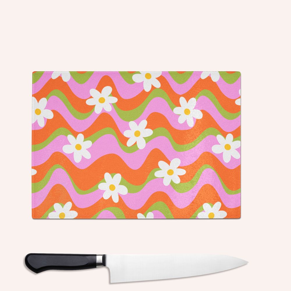 The Yililo Colourful Wave Flower Glass Chopping Board is a rectangular tempered glass kitchenware item with wavy orange, pink, and green patterns and white daisies. A large kitchen knife with a black handle is placed below it.
