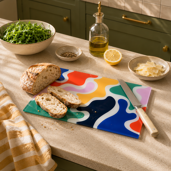 Colourful wavy camouflage style cutting board in primary colours with bread, knife, and ingredients on a kitchen counter.