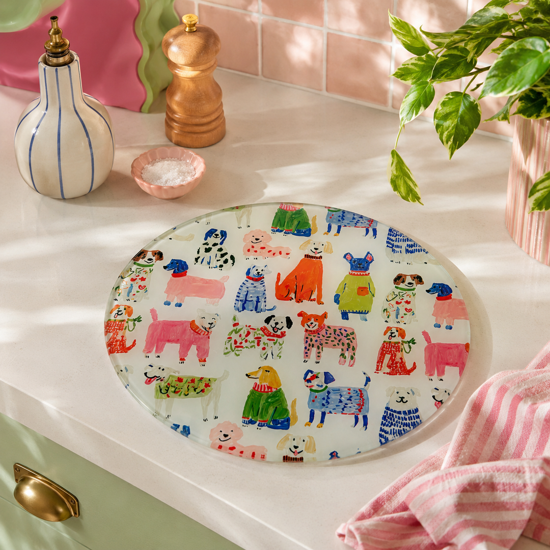 A round glass serving platter with a colorful cartoon dog pattern, styled on a white countertop with kitchen props like a striped bottle, a wooden pepper mill, and a pink striped towel.