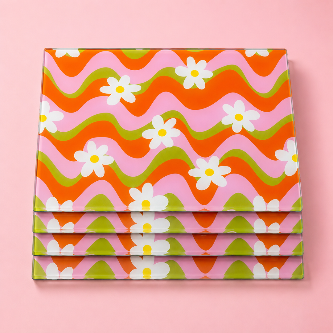 A stack of four rectangle glass placemats with a retro 1970s-style wavy pattern in orange, pink, and green, decorated with white daisy flowers, set against a solid pink background.