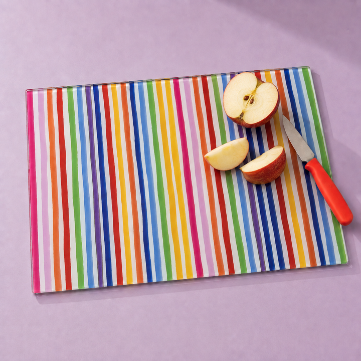A rectangular glass cutting board with a vibrant multicolor vertical stripe pattern, shown with sliced red apples and a small knife on a solid lavender background.