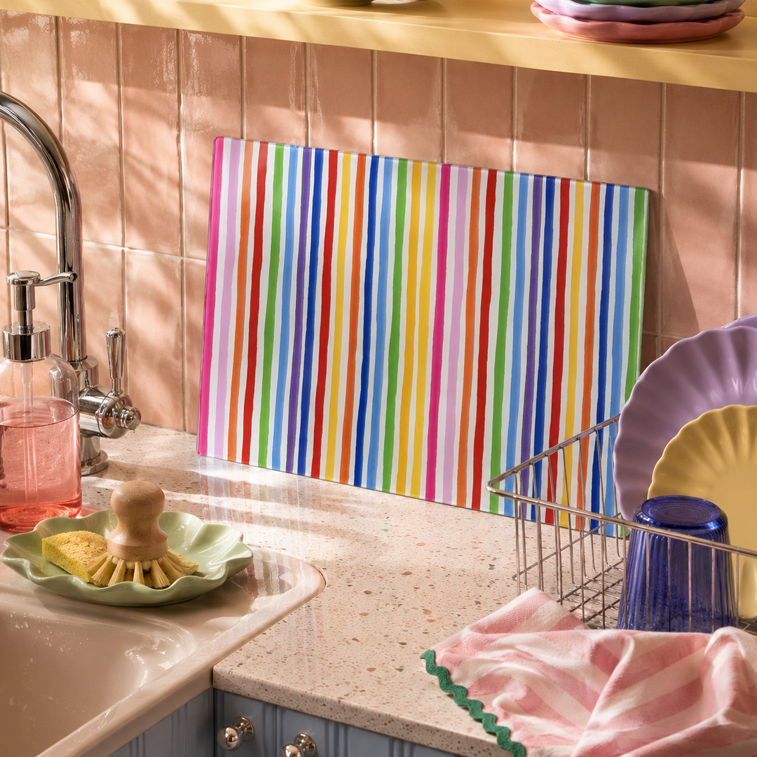 A colourful striped cutting board with vertical multicolour stripes leans against a peach tiled kitchen backsplash on a speckled countertop, surrounded by kitchen items like a sink, dish rack, and dishes.