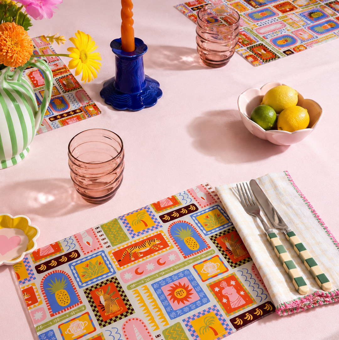 Colourful tropical table setting with glass placemats, cutlery, and a bowl of fruit on a white table.