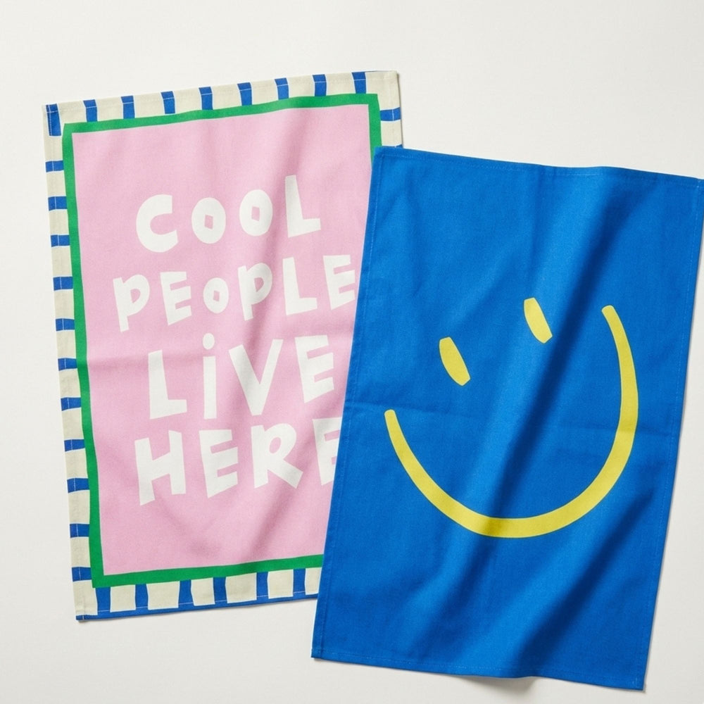 Two towels, one pink with 'Cool People Live Here' text and the other blue with a smiley face, on a light gray background.