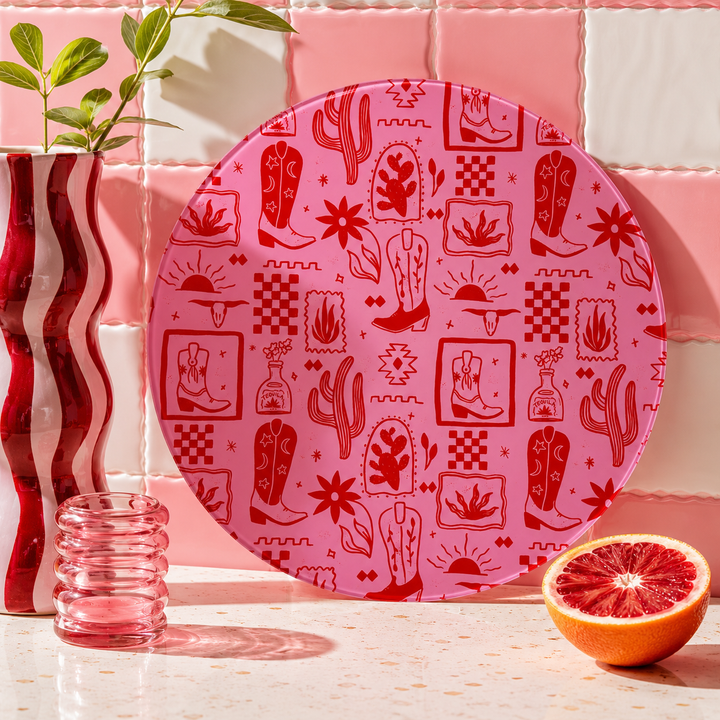 Pink decorative cowboy cowgirl round glass worktop saver with red patterns on a pink and white checkered background with a glass and half a grapefruit.