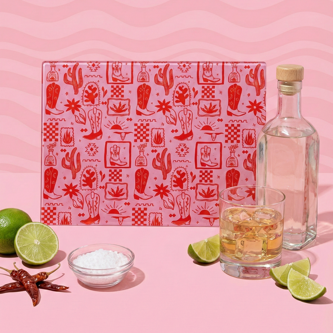 Tequila setup with a red patterned glass chopping board, glass of tequila, bottle, limes, and salt on a pink background