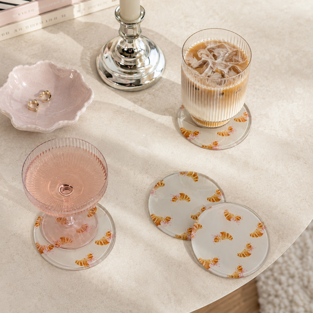 A set of 4 round glass croissant decorated coasters with coquette cute bows, with a ice coffee and wine glass on top placed on a cream table 