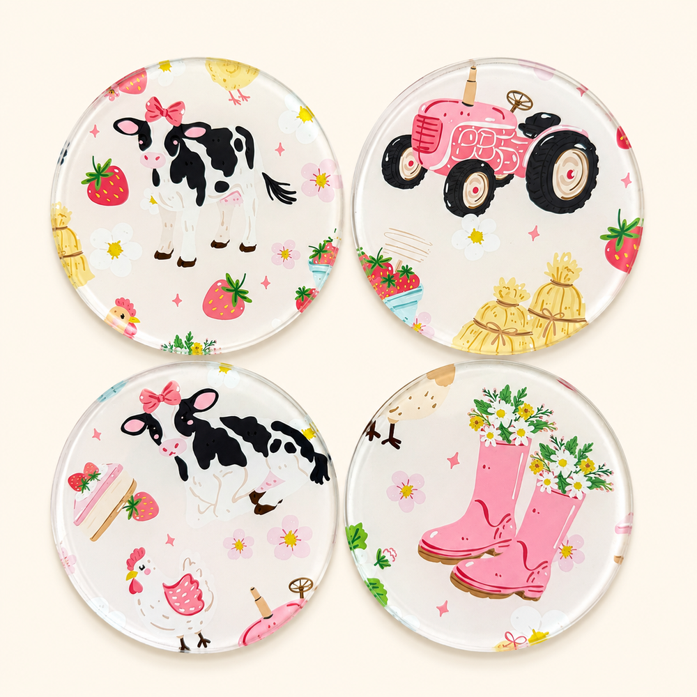 Four round glass 
clear coasters arranged in a 2x2 grid on a light cream background. Each coaster features a colorful farm-themed illustration, including black-and-white cows, pink tractors, strawberries, pink rain boots with flowers, chickens, and hay bales.