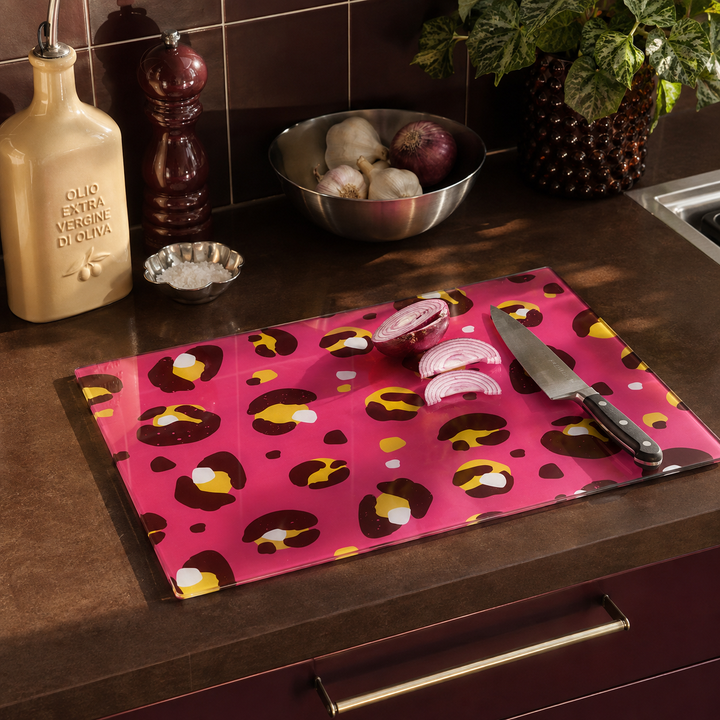 A rectangular glass cutting board with a pink leopard print pattern, placed on a dark kitchen countertop. The board is styled with a knife, sliced red onions, garlic, and kitchen accessories in the background.