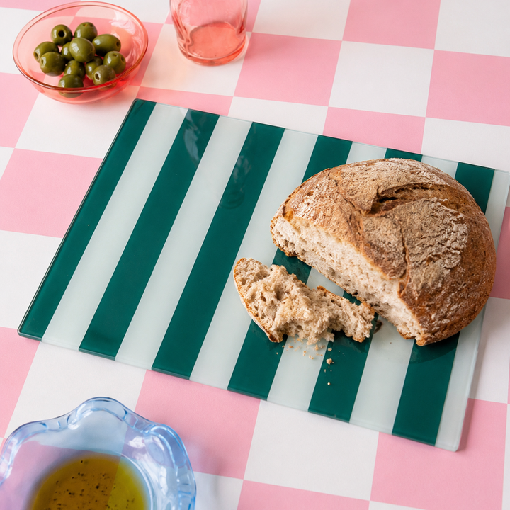 Loaf of bread with a slice cut off on a green striped glass cutting board with olives and oil on a pink checkered tablecloth.