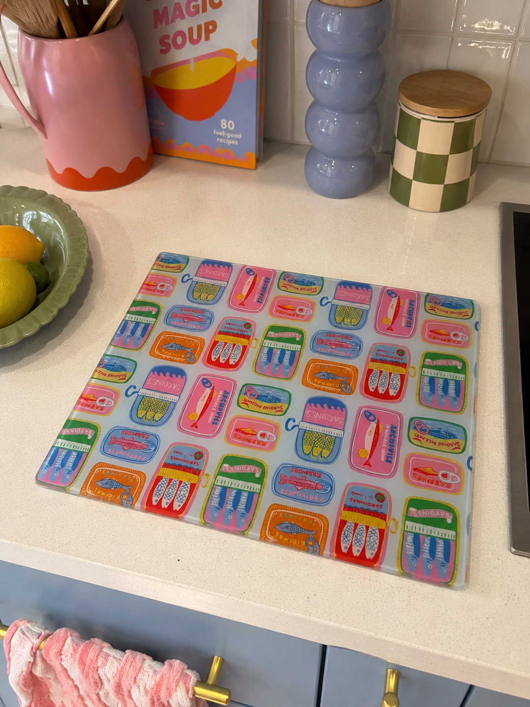 Colourful glass sardine parade glass worktop saver on a kitchen counter with various kitchen items in the background.
