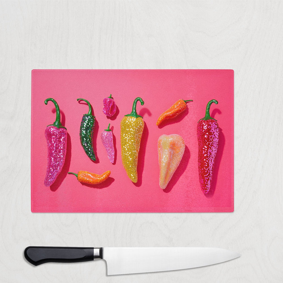 A row of colorful chili peppers covered in glitter is arranged on a Yililo Glitter Chilli Colourful Glass Chopping Board, with a chef’s knife placed below on a white surface.
