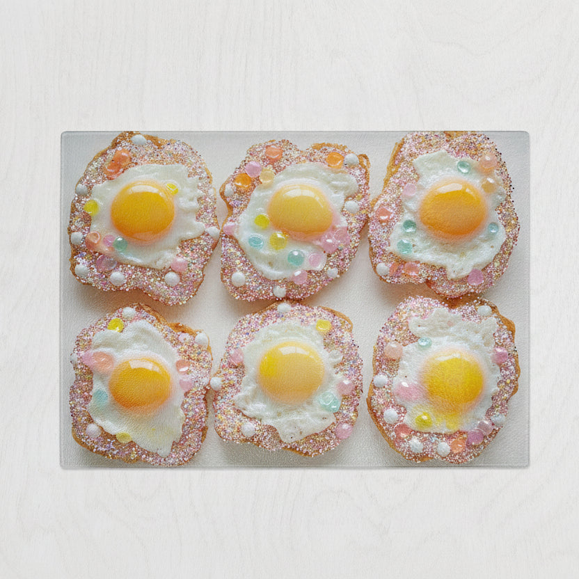 Six pieces of toast with candies and toy eggs are arranged on a pastel Yililo Glitter Egg Glass Chopping Board, with a chef’s knife placed below on a light-colored table.
