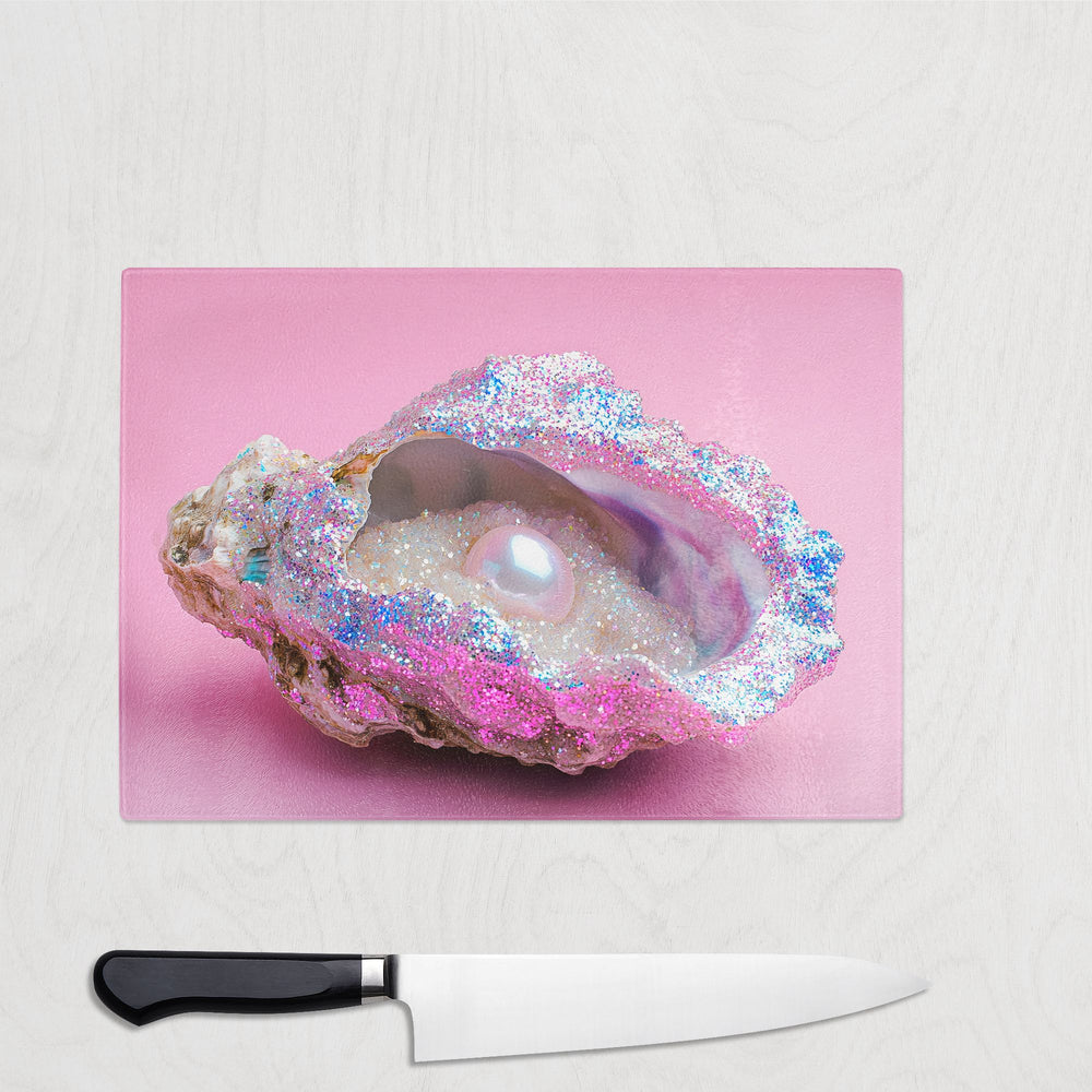 An iridescent oyster shell with a glittering pearl and shimmer sits on pink. Below, a chef’s knife rests on the Yililo Glitter Pearl Oyster Pink Glass Chopping Board—perfect for stylish kitchen prep.