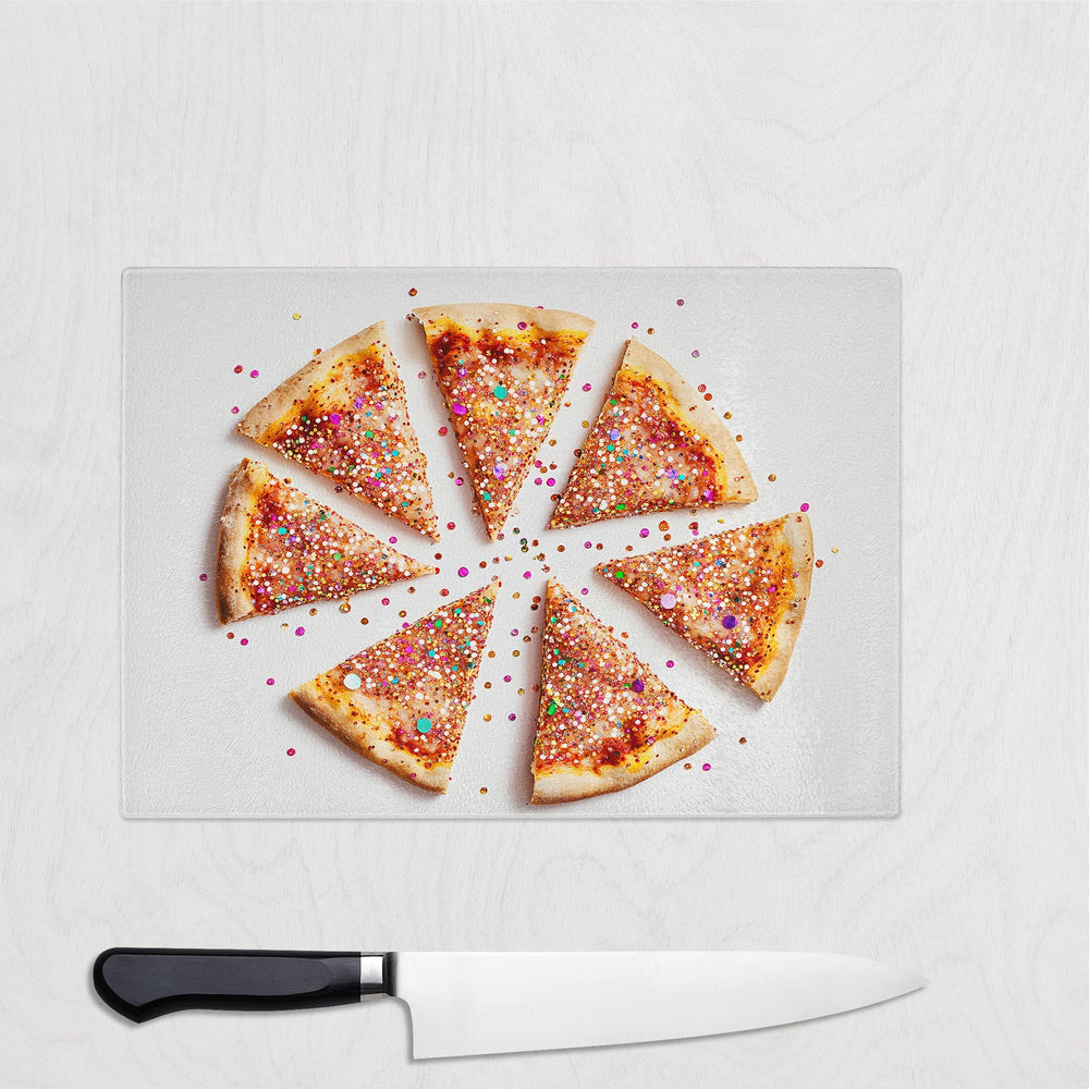 Seven slices of glitter pizza with colorful sprinkles are arranged in a circle on the Yililo Glitter Pizza Glass Chopping Board, with a chef’s knife below on a light wooden surface.