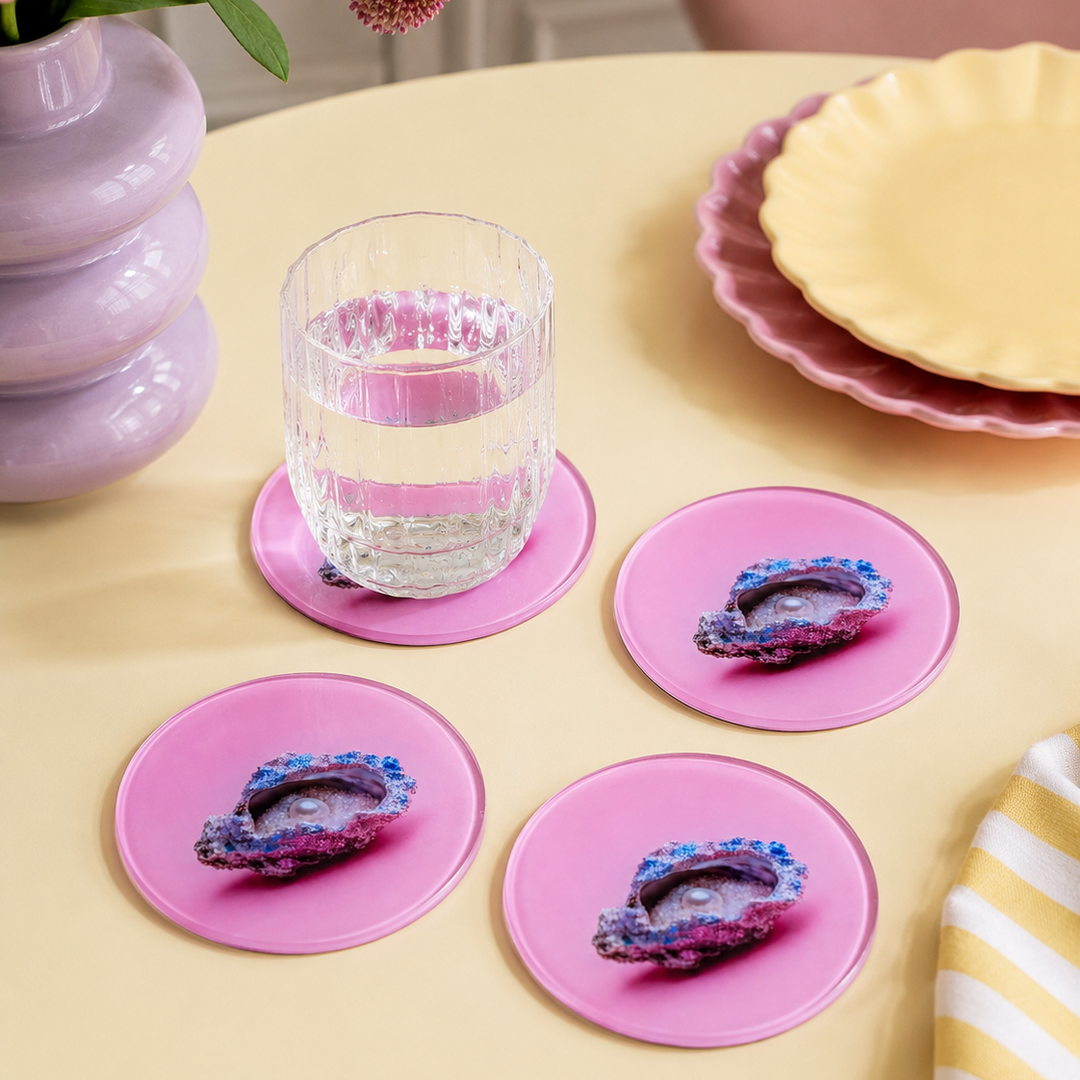 Four round pink coasters featuring geode designs, arranged on a pale yellow table with a glass of water on one coaster. A lavender vase with flowers, stacked pink and yellow plates, and a striped cloth are visible in the styled setting.