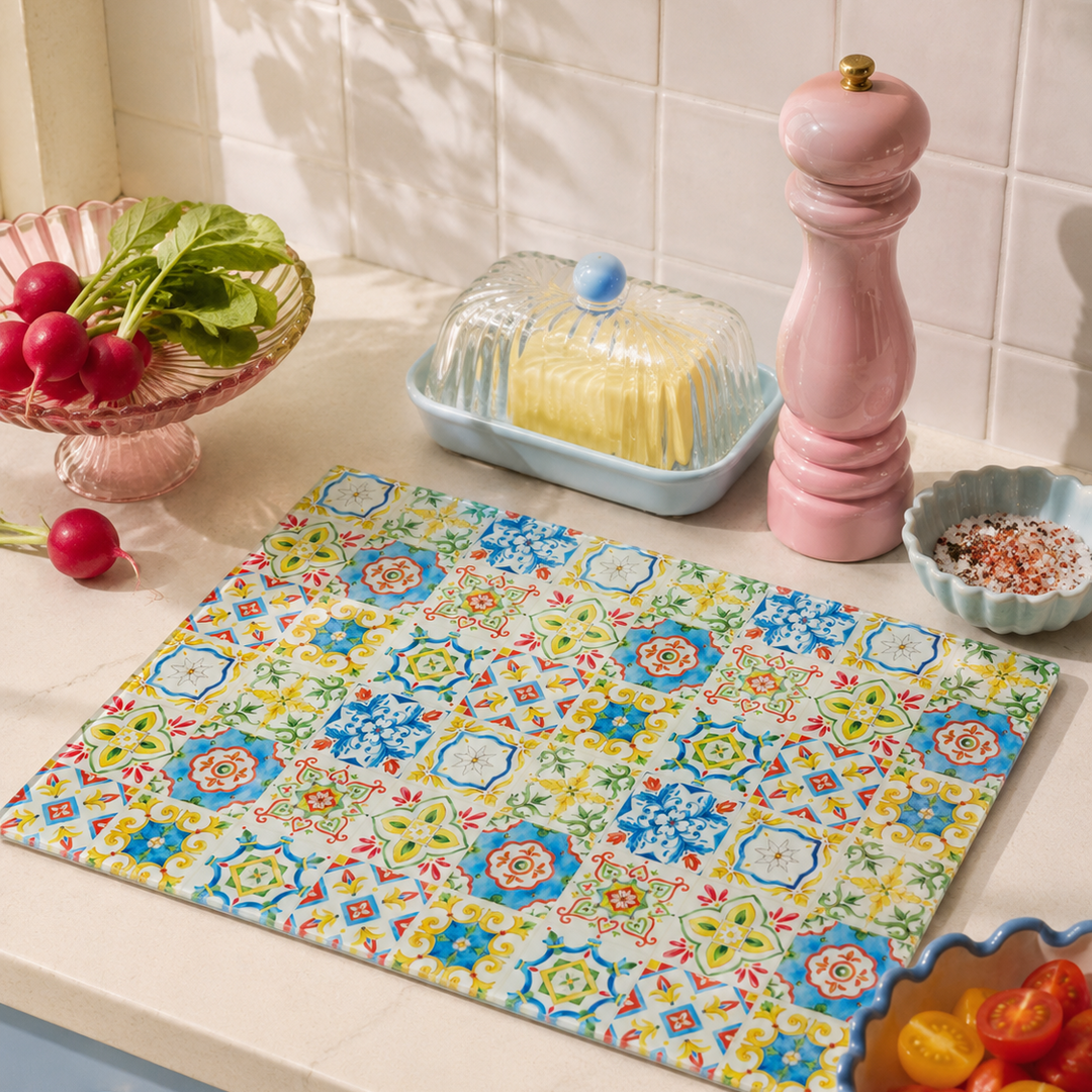 A rectangular kitchen glass chopping board mat with a traditional colourful  Mediterranean-style tile pattern in blue, yellow, green, and red, laid out on a cream countertop in a kitchen setting.