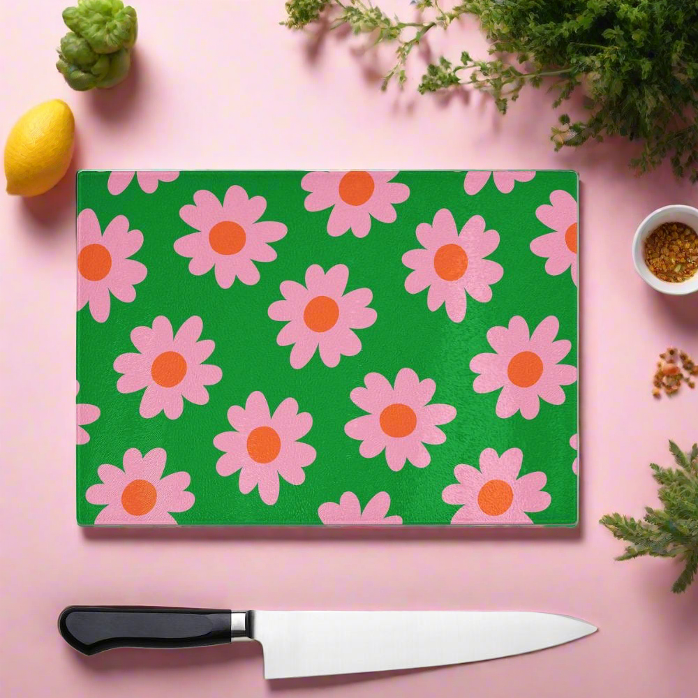 Brighten your kitchen with the Yililo Green And Pink Flower Glass Chopping Board, featuring a durable green tempered glass surface with a pink daisy pattern—captured in a setting of herbs, lemon, spices, and knife on pink background.