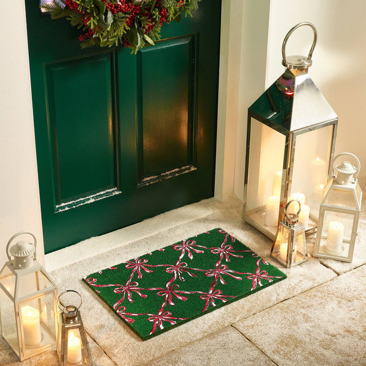 Decorative doormat with pink bows in front of a green door with festive wreath, surrounded by lanterns and candles.