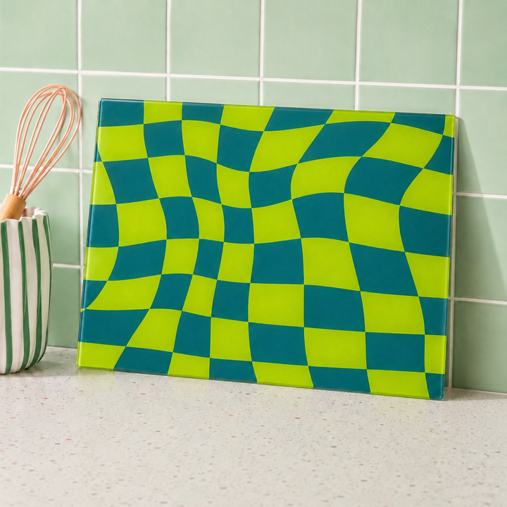 Green and blue checkered glass cutting board on a kitchen counter with a whisk in the background.