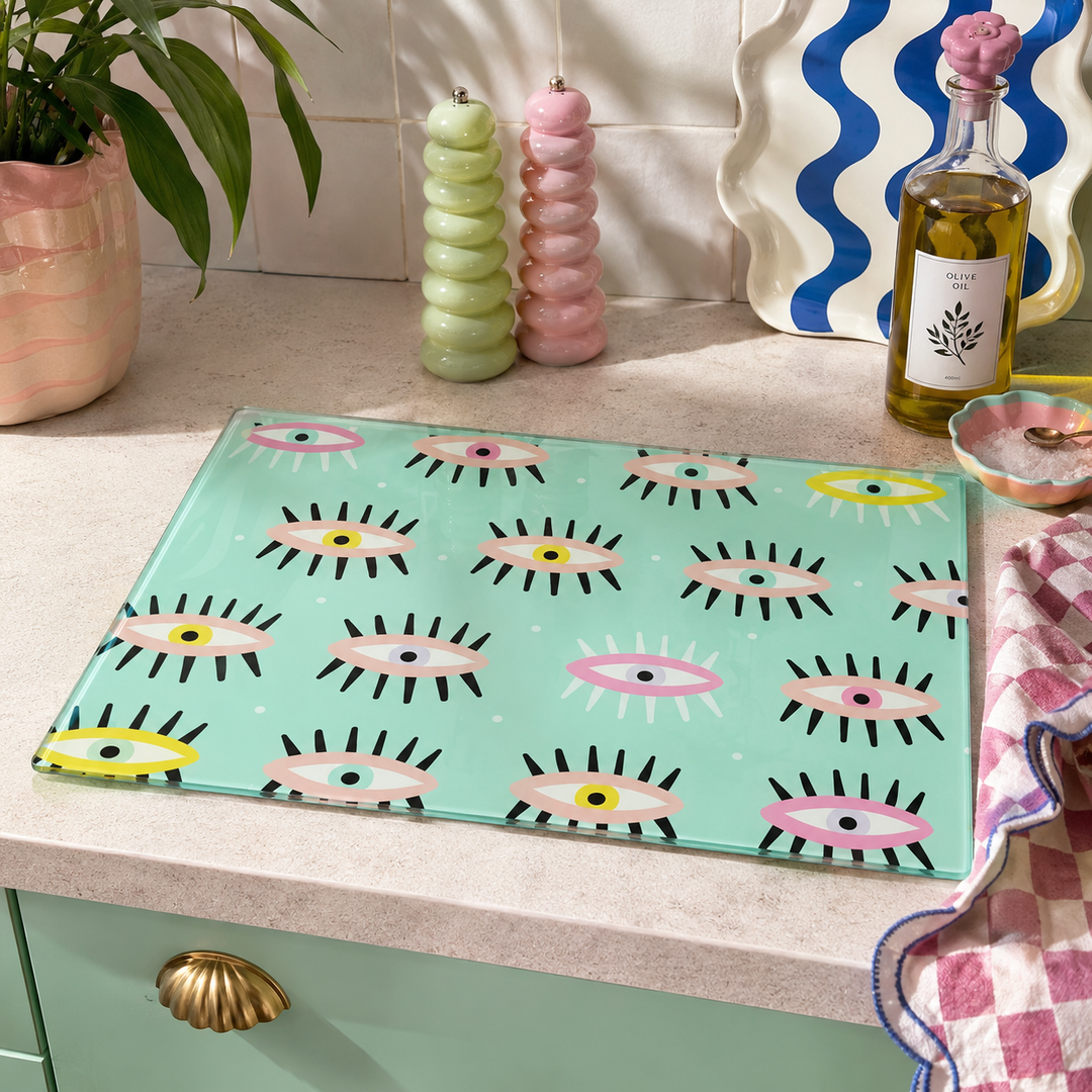 A rectangular glass cutting board with a repeating pattern of stylized pink, yellow, and black eyes on a mint green background, placed on a kitchen counter next to a pink-and-white checkered cloth, candles, and a bottle of oil.